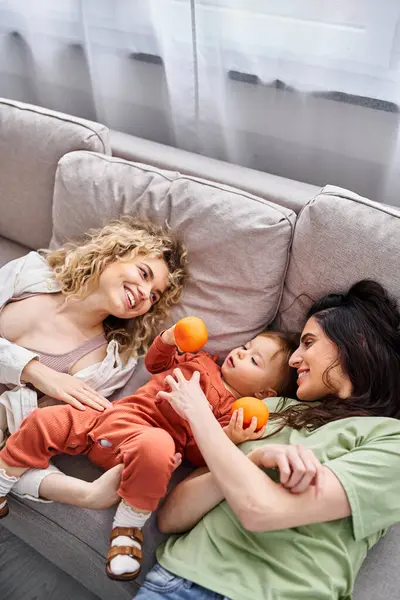 Joyful lgbt couple lying on sofa with their pretty baby girl holding tangerines, family concept — Stock Photo
