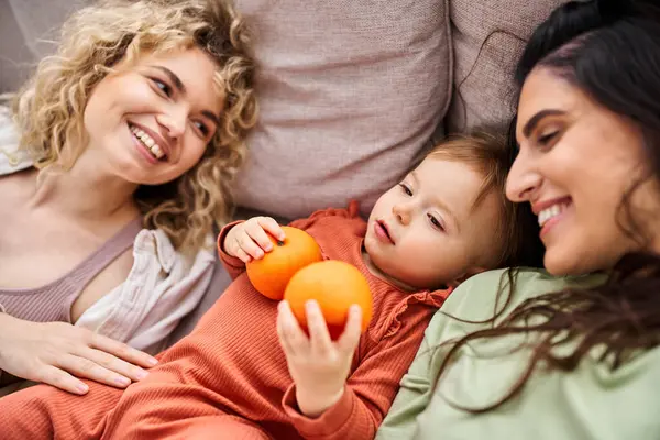 Merry lgbt couple lying on sofa with their pretty baby girl holding tangerines, family concept — Stock Photo