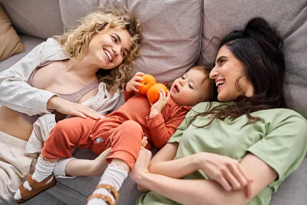 Happy lgbt couple lying on sofa with their pretty baby girl holding tangerines, family concept — Stock Photo