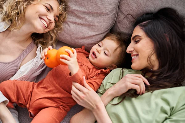 Beautiful jolly lgbt couple lying on sofa with their baby girl holding tangerines, family concept — Stock Photo