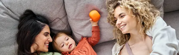 Joyeux couple lgbt couché sur le canapé avec leur jolie petite fille tenant des mandarines, concept de famille, bannière — Photo de stock