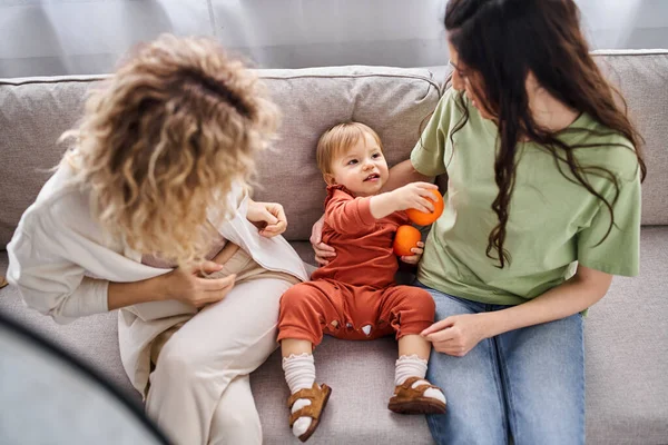 Fürsorgliche lustige lgbt Paar sitzt auf dem Sofa mit ihrem kleinen Mädchen mit Mandarinen, Familienkonzept — Stockfoto