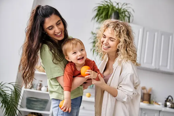 Joyful lesbian couple having fun together with their cute baby girl holding tangerines, family — Stock Photo