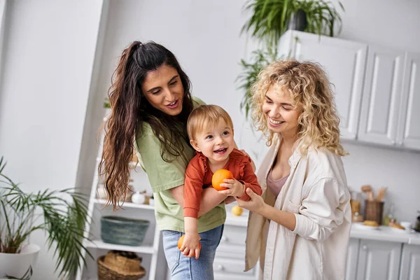 Joyous lesbian couple having fun together with their cute baby girl holding tangerines, family — Stock Photo