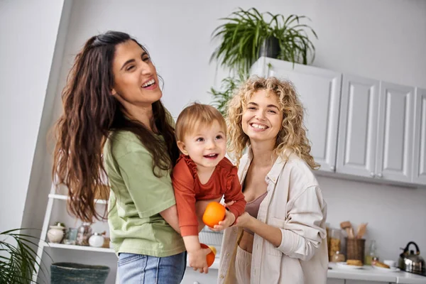 Jolly lesbian couple having fun together with their cute baby girl holding tangerines, family — Stock Photo