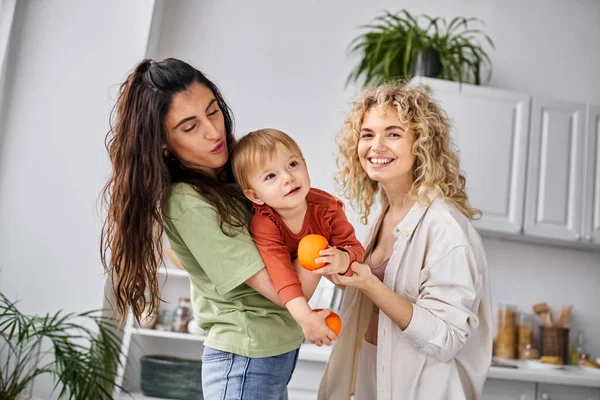 Feliz casal muito lgbt se divertindo junto com sua linda menina segurando tangerinas, família — Fotografia de Stock