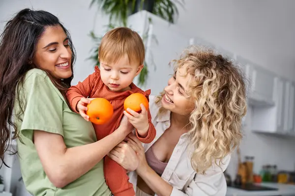 Loving lgbt couple having fun together with their cute baby girl holding tangerines, family concept — Stock Photo
