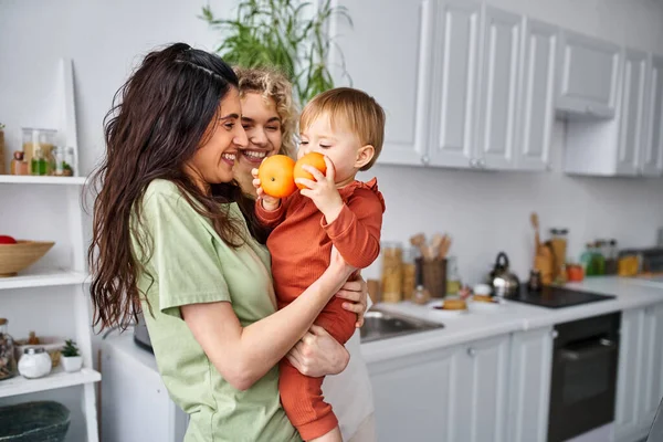 Attractive lgbt couple having fun together with their cute baby girl holding tangerines, family — Stock Photo