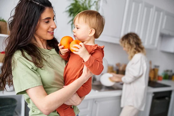 Fokus auf hübsche Frau mit Tochter, die Mandarinen mit ihrem verschwommenen blonden Partner vor dem Hintergrund hält — Stockfoto