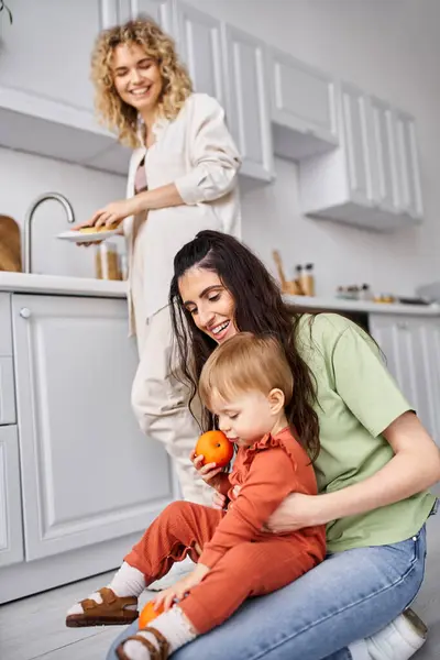 Focus on cheerful woman with daughter holding tangerines with her blurred blonde partner on backdrop — Stock Photo