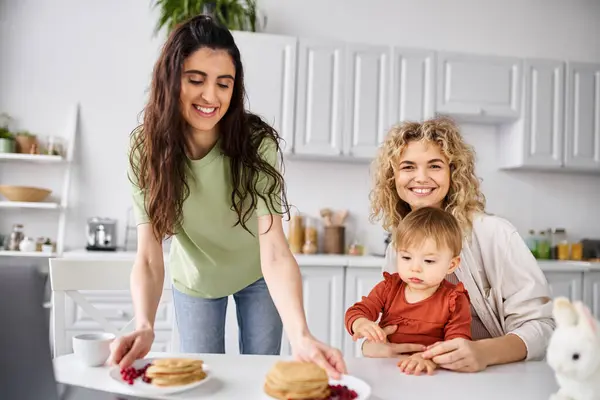 Atractivo alegre pareja lesbiana teniendo panqueques en el desayuno con su bebé niña, concepto de familia - foto de stock