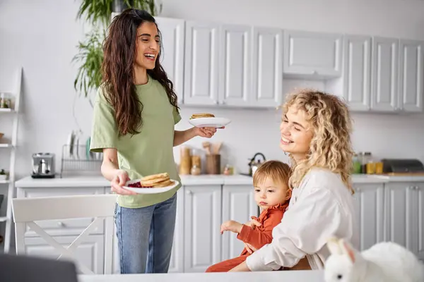 Attraktive blonde Frau bereitet ihrem verschwommenen Baby Pfannkuchen zum Frühstück zu, Familienkonzept — Stockfoto