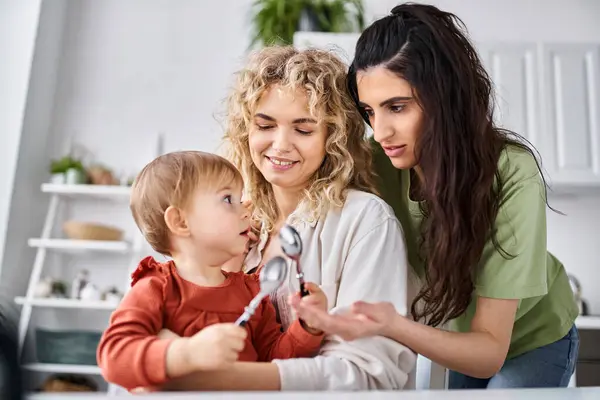 Charmant couple lgbt en vêtements de maison passer du temps avec leur fille sur la cuisine, cuillères à la main — Photo de stock