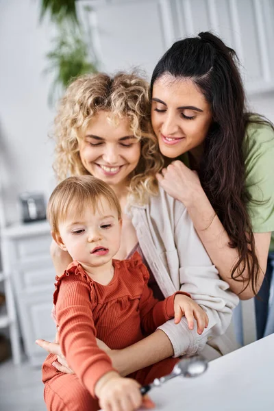 Attractive lgbt couple in homewear spending time with their daughter on kitchen, spoons in hand — Stock Photo
