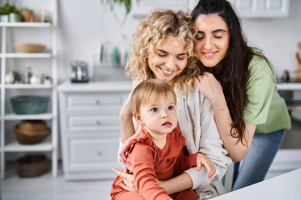 Joyful lgbt couple in homewear spending time with their daughter on kitchen, spoons in hand — Stock Photo