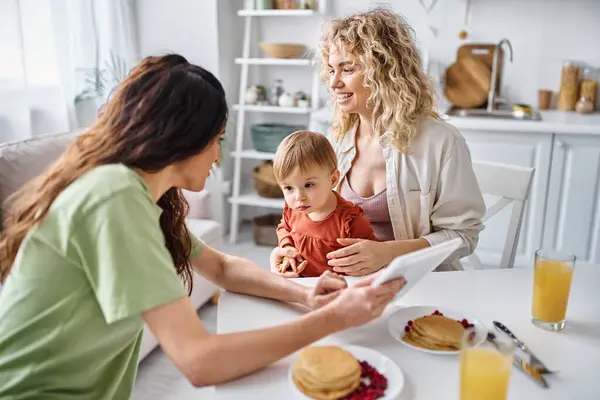 Attractive lgbt couple having breakfast with their baby girl and looking at tablet, family concept — Stock Photo