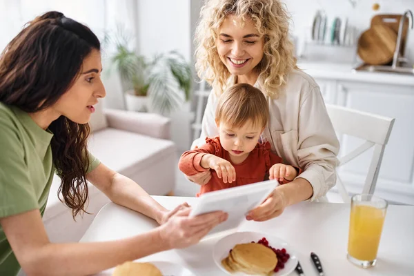 Beautiful lgbt couple having breakfast with their baby girl and looking at tablet, family concept — Stock Photo