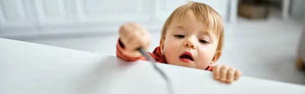 Linda niña en suéter naranja acogedor tratando de llegar a un poco de desayuno en la mesa, pancarta - foto de stock