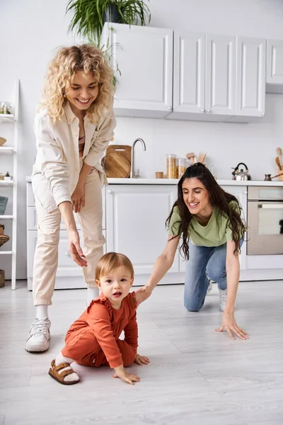 Joyous lesbian couple in homewear looking how their cute baby girl crawling, family concept — Stock Photo