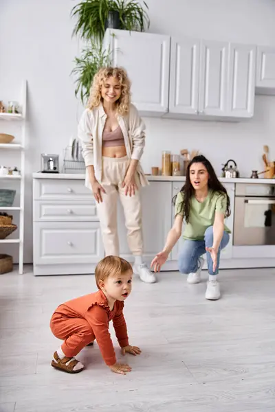 Cheerful lesbian couple in homewear looking how their cute baby girl crawling, family concept — Stock Photo