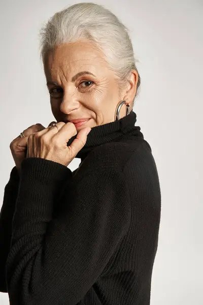 Portrait of pleased middle aged woman adjusting collar of turtleneck sweater on grey backdrop — Stock Photo