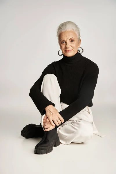 Mujer de negocios feliz de mediana edad en elegante atuendo casual elegante sentado sobre fondo gris - foto de stock