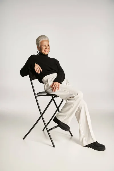 Glückliche Geschäftsfrau mittleren Alters in schicker Freizeitkleidung und Reifrohren, die auf einem Stuhl auf grau sitzt — Stockfoto