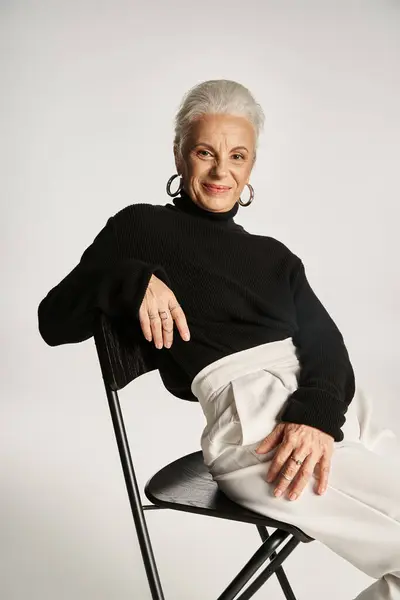 Alegre mujer de negocios de mediana edad en traje casual inteligente y pendientes de aro sentado en la silla en gris - foto de stock
