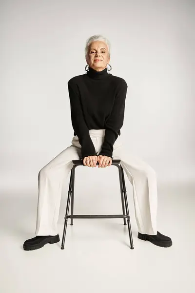 Elegante mujer de negocios de mediana edad en traje casual inteligente y pendientes de aro sentado en la silla en gris - foto de stock
