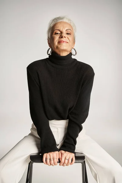 Soddisfatto donna d'affari di mezza età in abbigliamento casual intelligente e orecchini a cerchio seduto sulla sedia su grigio — Foto stock