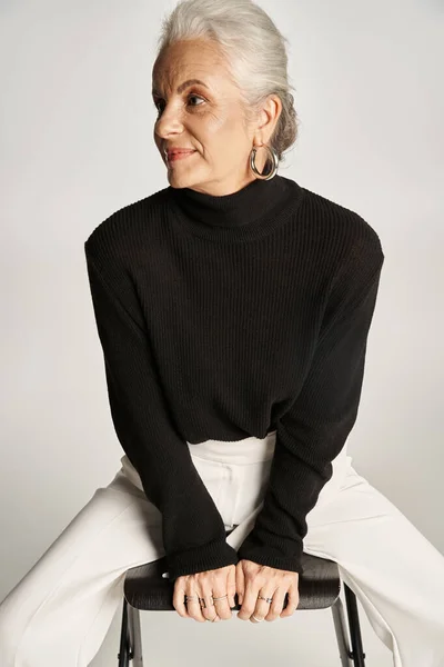 Zufriedene Geschäftsfrau mittleren Alters in schicker Freizeitkleidung und Reifrohren sitzt auf einem Stuhl auf grau — Stockfoto