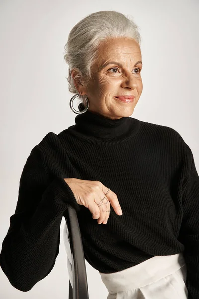 Charming middle aged woman in smart casual attire and hoop earrings sitting on chair on grey — Stock Photo