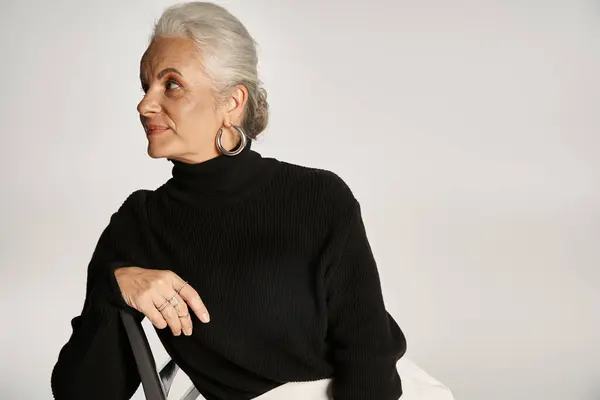 Encantadora mujer de mediana edad en traje casual inteligente y pendientes de aro sentado en la silla en gris - foto de stock
