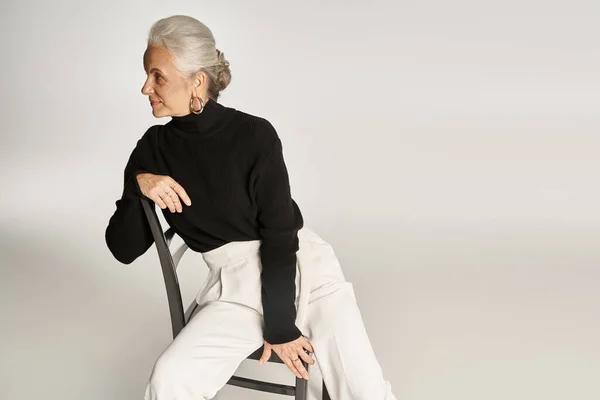 Attraente donna di mezza età in elegante abbigliamento casual e orecchini a cerchio seduto sulla sedia su grigio — Foto stock