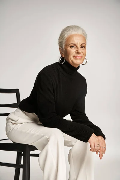 Attraente donna di mezza età in elegante abbigliamento casual e orecchini a cerchio seduto sulla sedia su grigio — Foto stock