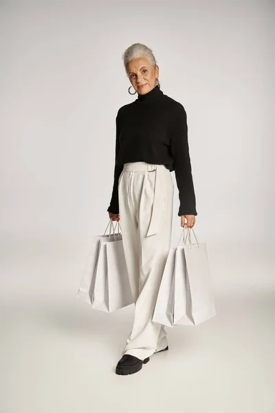 Felice donna di mezza età in elegante abbigliamento e orecchini a cerchio in piedi con borse della spesa su grigio — Foto stock