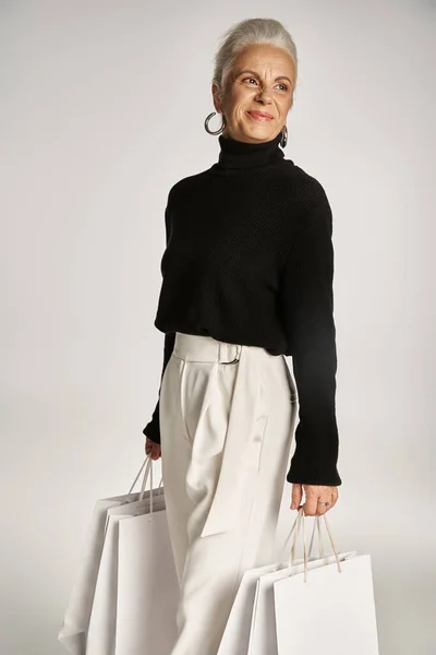 Fröhliche Frau mittleren Alters in eleganter Kleidung und Reifrohrringen stehend mit Einkaufstaschen auf grau — Stockfoto