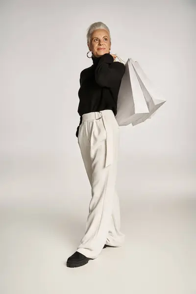 Verträumte Frau mittleren Alters in eleganter Kleidung und Reifrohrringen mit Einkaufstaschen auf grau — Stockfoto
