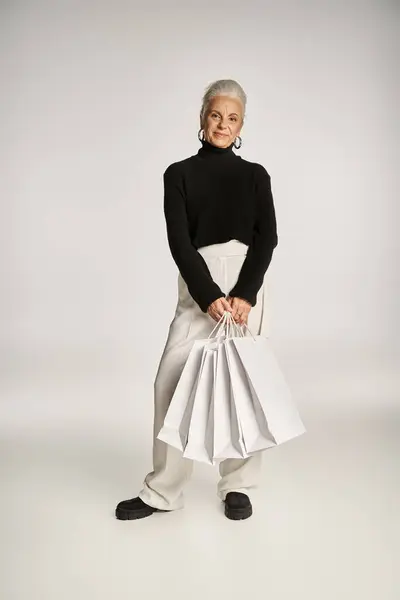 Zufriedene Frau mittleren Alters in eleganter Kleidung und Reifrohrringen mit Einkaufstaschen auf grau — Stockfoto