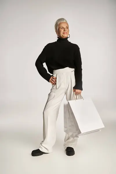 Soddisfatta donna di mezza età in elegante abbigliamento e orecchini a cerchio in piedi con borse della spesa su grigio — Foto stock