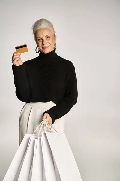 Donna di mezza età soddisfatta in abbigliamento elegante con carta di credito e borse della spesa su sfondo grigio — Foto stock