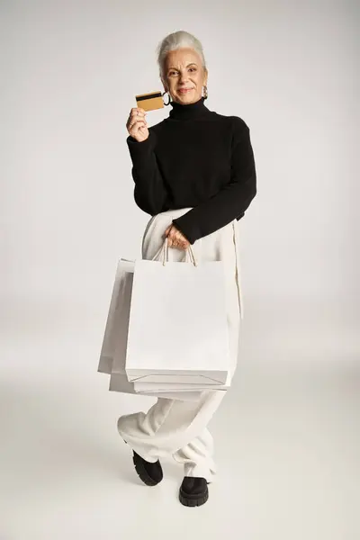 Satisfied middle aged woman in elegant attire holding credit card and shopping bags on grey backdrop — Stock Photo