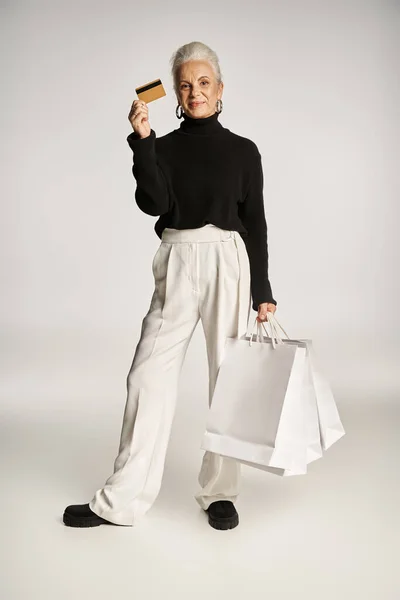 Donna di mezza età soddisfatta in abbigliamento elegante con carta di credito e borse della spesa su sfondo grigio — Foto stock