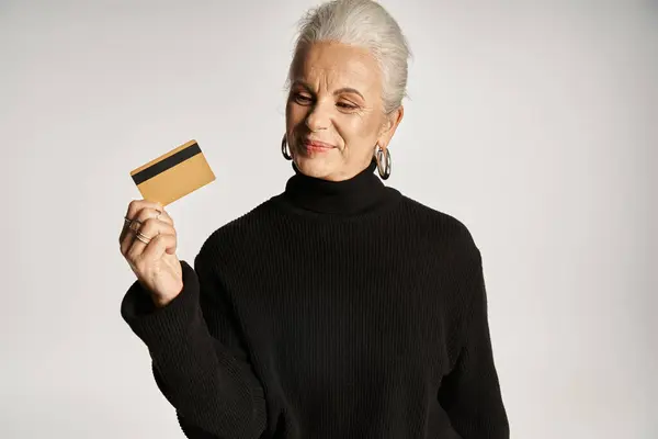 Elegante donna di mezza età in maglione dolcevita nero con carta di credito su sfondo grigio — Foto stock