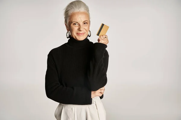 Elegant middle aged woman in black turtleneck sweater holding credit card on grey backdrop — Stock Photo