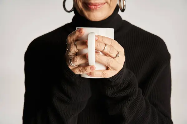 Cropped middle aged woman in black turtleneck sweater holding cup with hot beverage on grey backdrop — Stock Photo