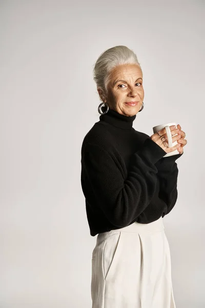 Pleased middle aged woman in black turtleneck sweater holding cup of coffee on grey backdrop — Stock Photo