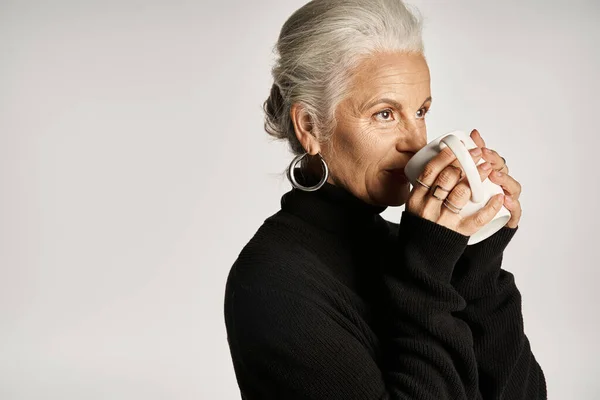 Happy middle aged woman in black turtleneck sweater holding cup of coffee on grey backdrop — Stock Photo