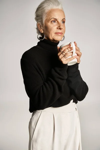 Charming middle aged woman in black turtleneck sweater holding cup with coffee on grey backdrop — Stock Photo