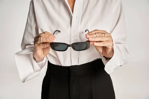 Tiro recortado de mujer de mediana edad en camisa blanca sosteniendo gafas de sol y de pie sobre fondo gris - foto de stock
